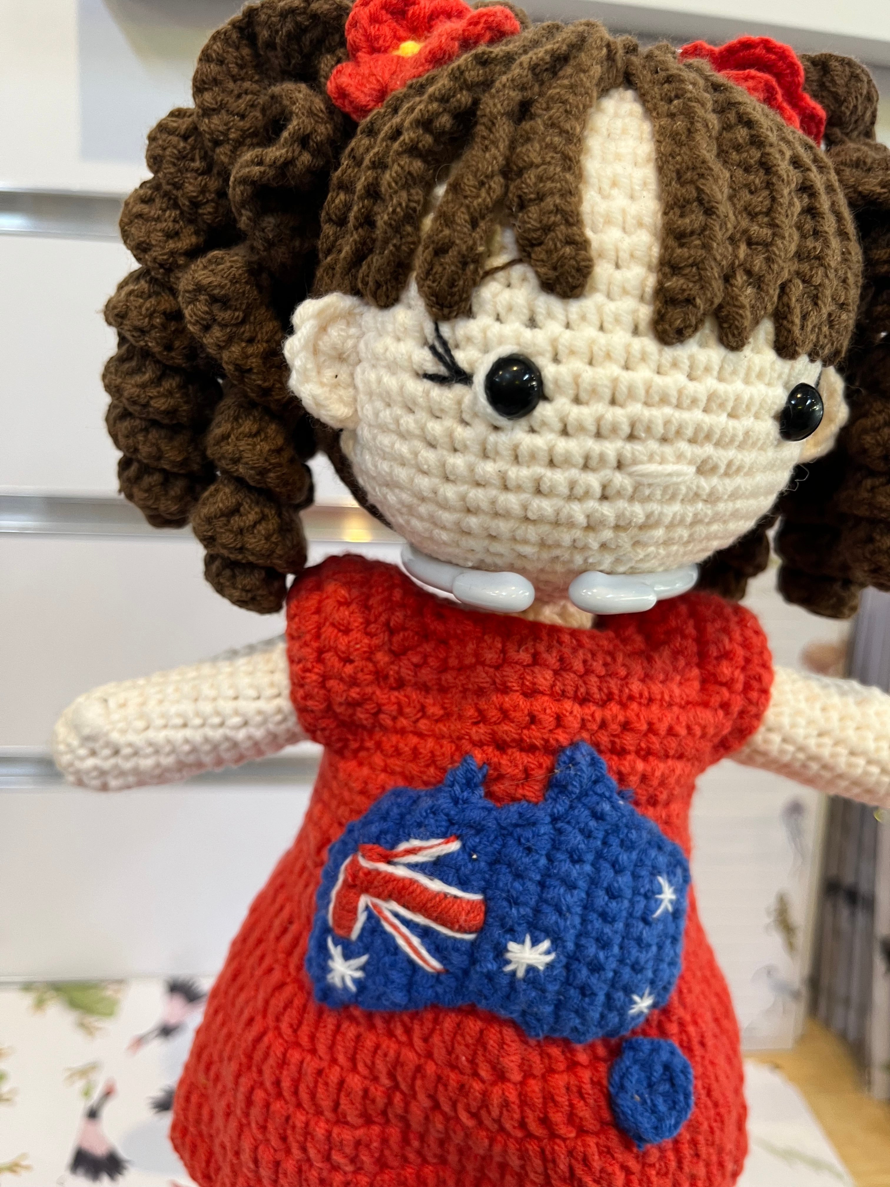 Suzie's Sunshine Dolls Mia, a crocheted doll with long brown curly hair, wearing a red dress with an Australian flag map applique.