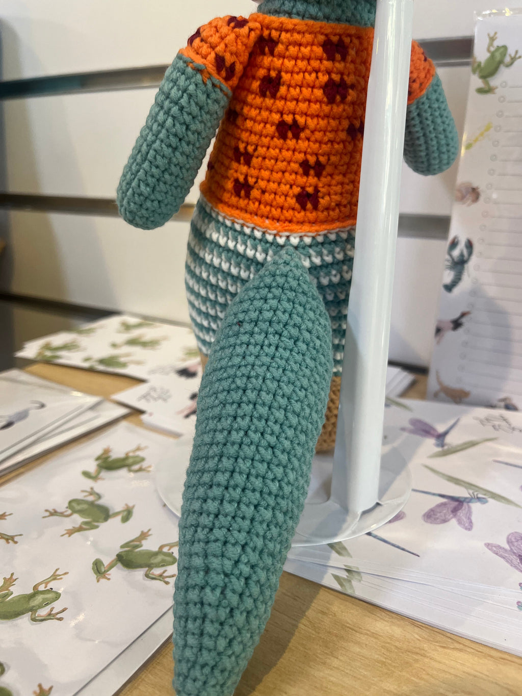 Suzie's Sunshine Dolls Calvin the Croc, a handmade crochet toy with a teal body, orange shirt with maroon paw prints, and striped pants.