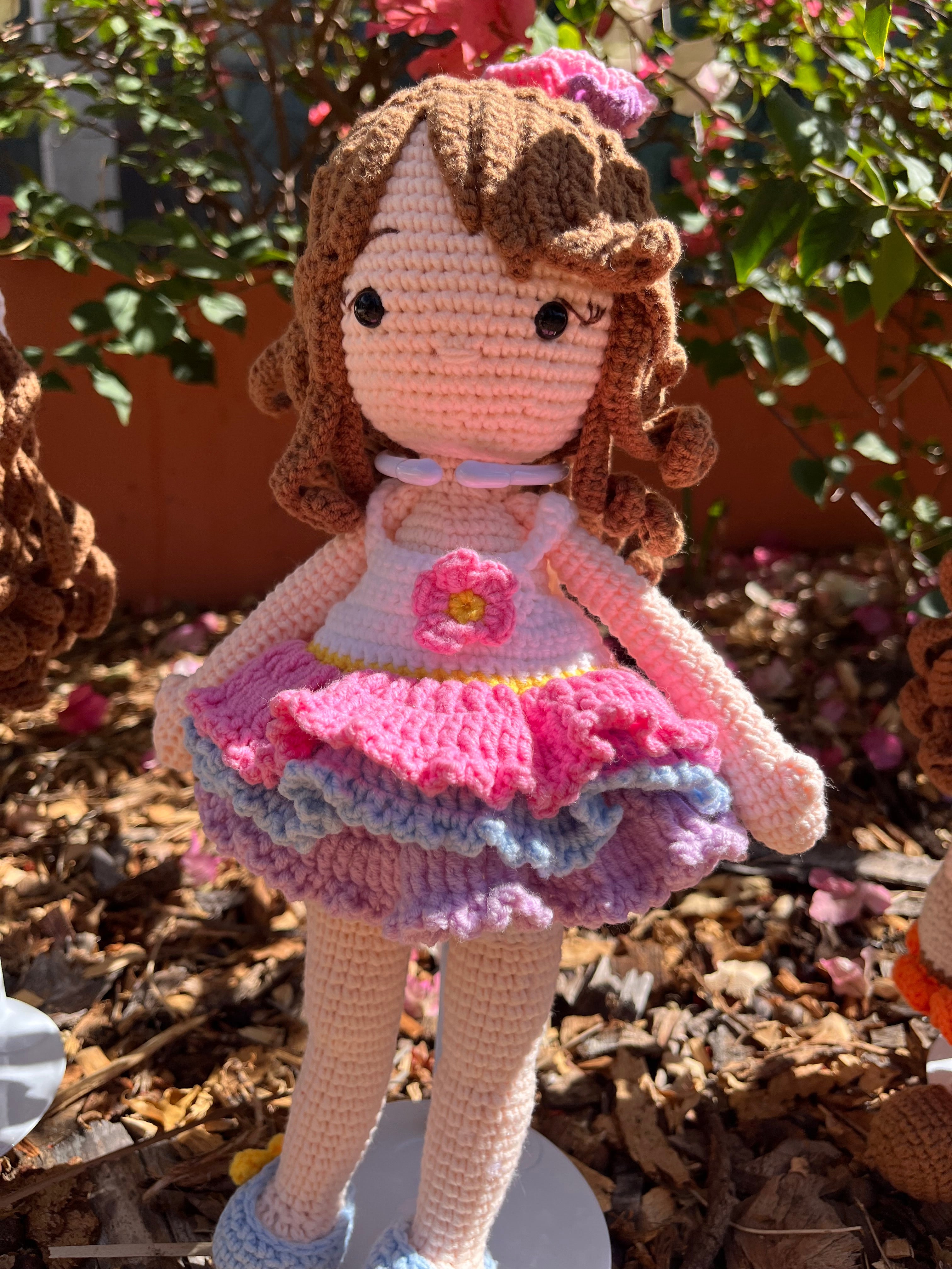 Suzie's Sunshine Dolls Cleo, a handmade crochet ballerina doll with brown curls, a pink, purple, and blue ruffled dress, and a flower applique.