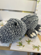 Suzie's Sunshine Dolls Kurtis the Croc, a handmade grey crochet crocodile plush toy with white toes and stitched smile.