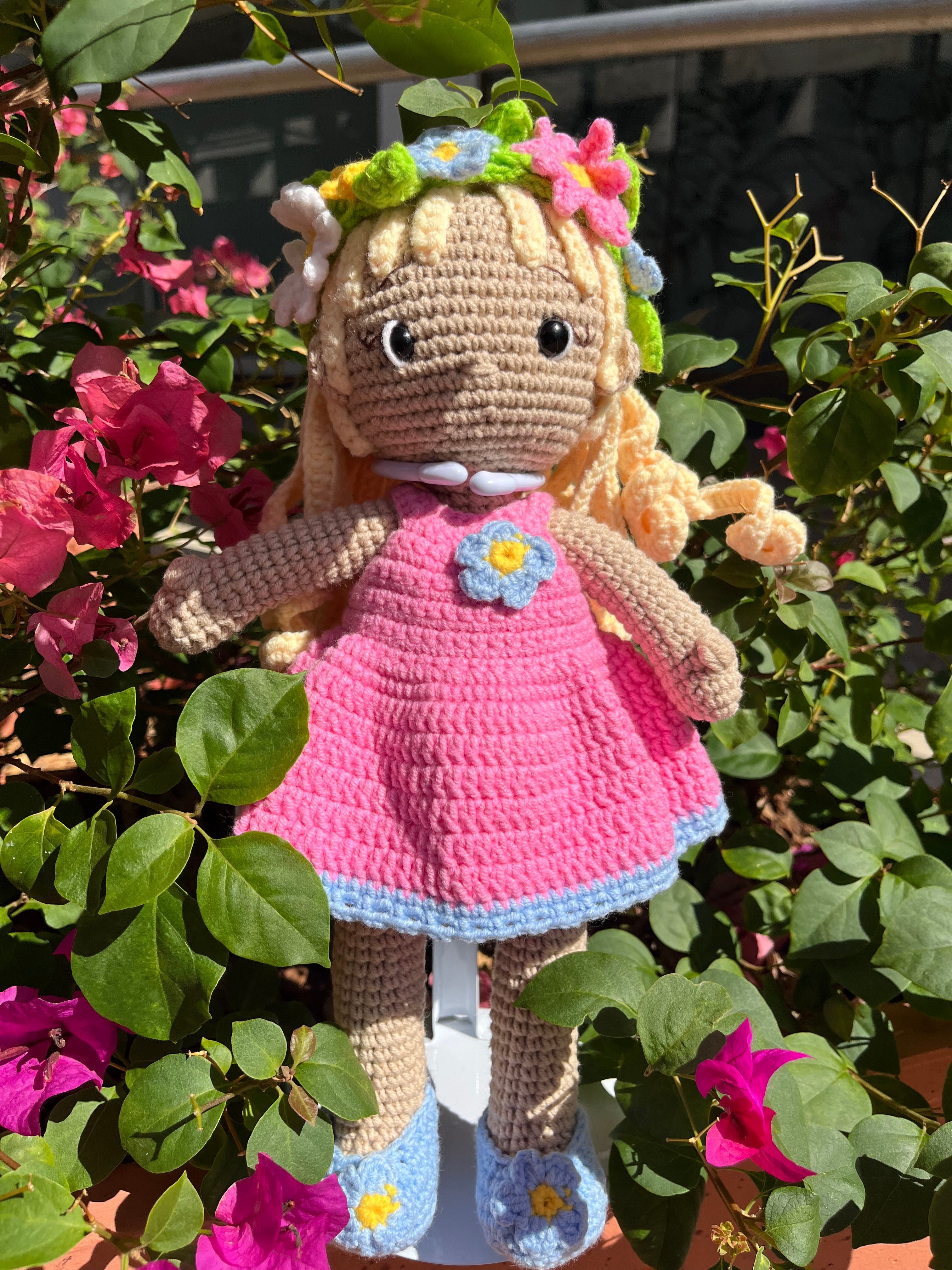 Suzie's Sunshine Dolls Delilah, a handmade crochet doll with blonde curls, wearing a pink dress with blue trim, blue shoes, and a floral crown.