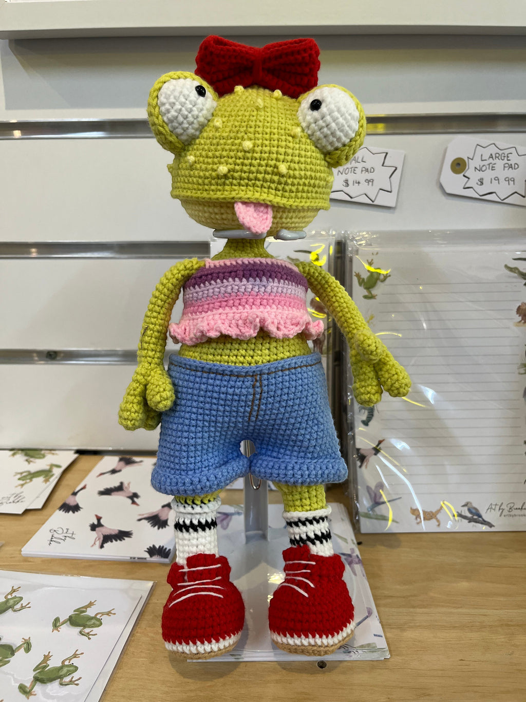 Suzie's Sunshine Dolls Frieda the Frog, a hand-crocheted doll with big googly eyes, red bow, and tongue out. She wears a pastel crop top, denim shorts, striped socks, and red sneakers.