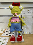Suzie's Sunshine Dolls Frieda the Frog, a hand-crocheted doll with big googly eyes, red bow, and tongue out. She wears a pastel crop top, denim shorts, striped socks, and red sneakers.