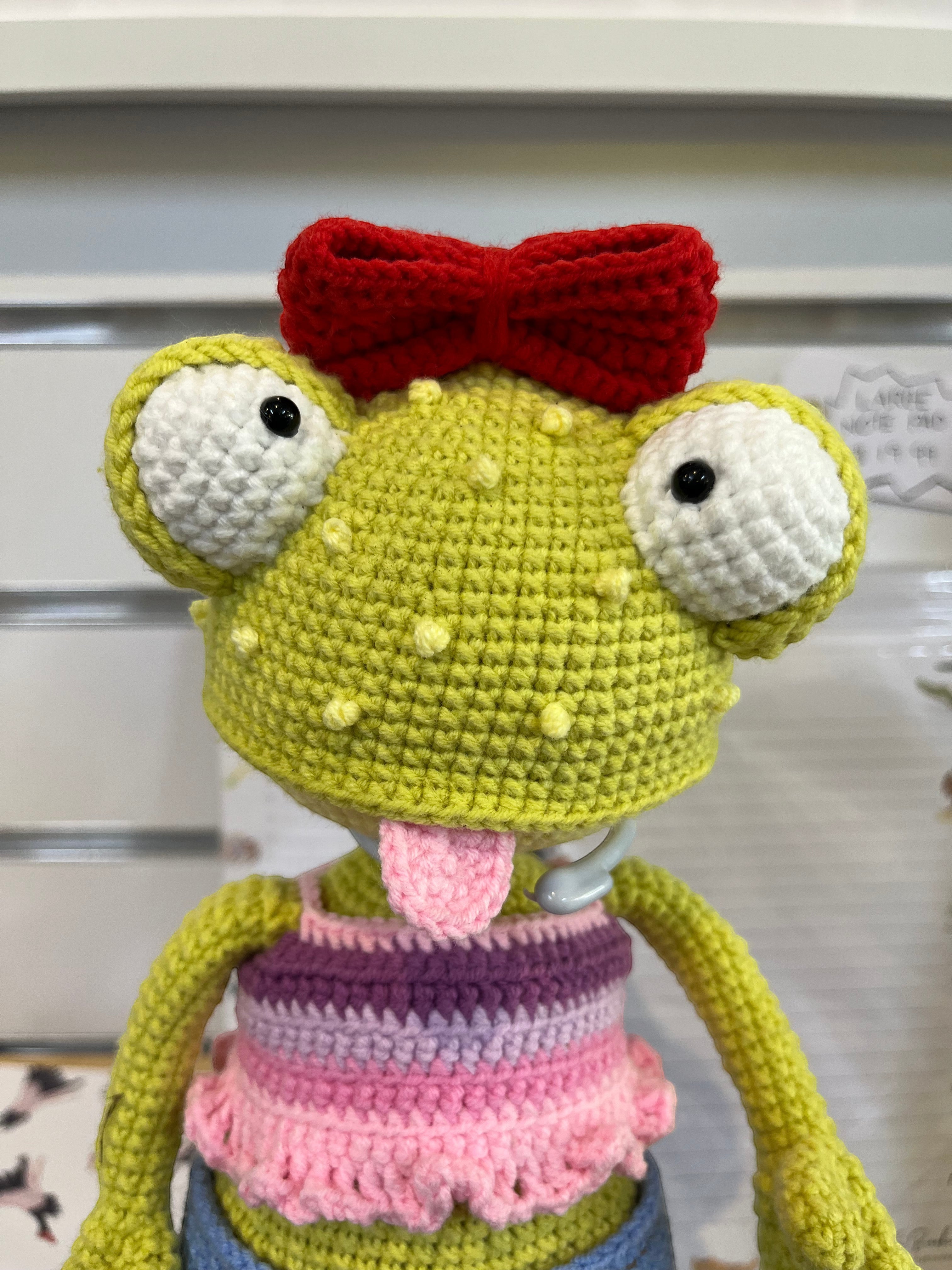 Suzie's Sunshine Dolls Frieda the Frog crochet doll with googly eyes, red bow, pink tongue out, and striped top.