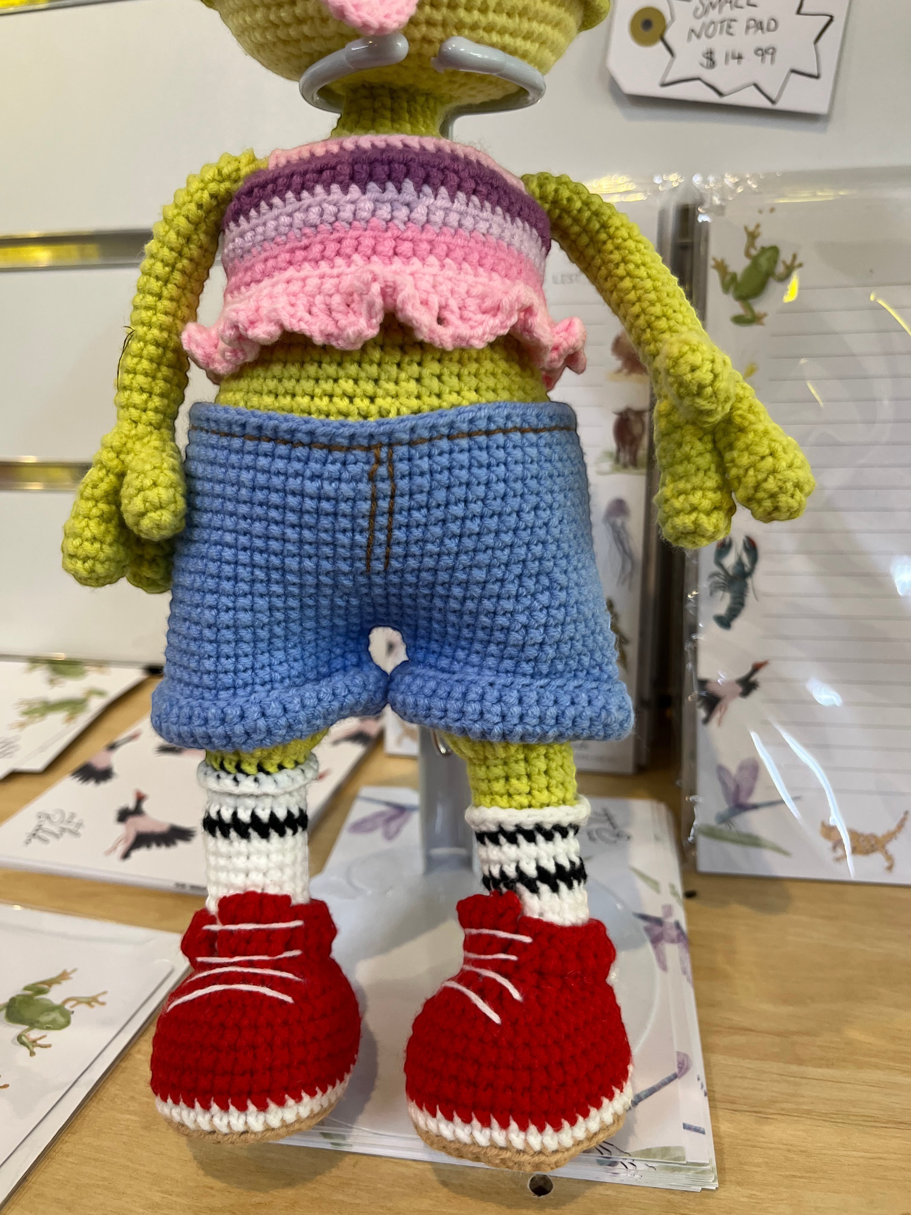 Suzie's Sunshine Dolls Frieda the Frog crochet doll with googly eyes, red bow, pastel crop top, denim shorts, striped socks, and red sneakers.
