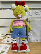 Suzie's Sunshine Dolls Frieda the Frog, a hand-crocheted doll with googly eyes, red bow, and tongue out. She wears a pastel crop top, denim shorts, striped socks, and red sneakers.