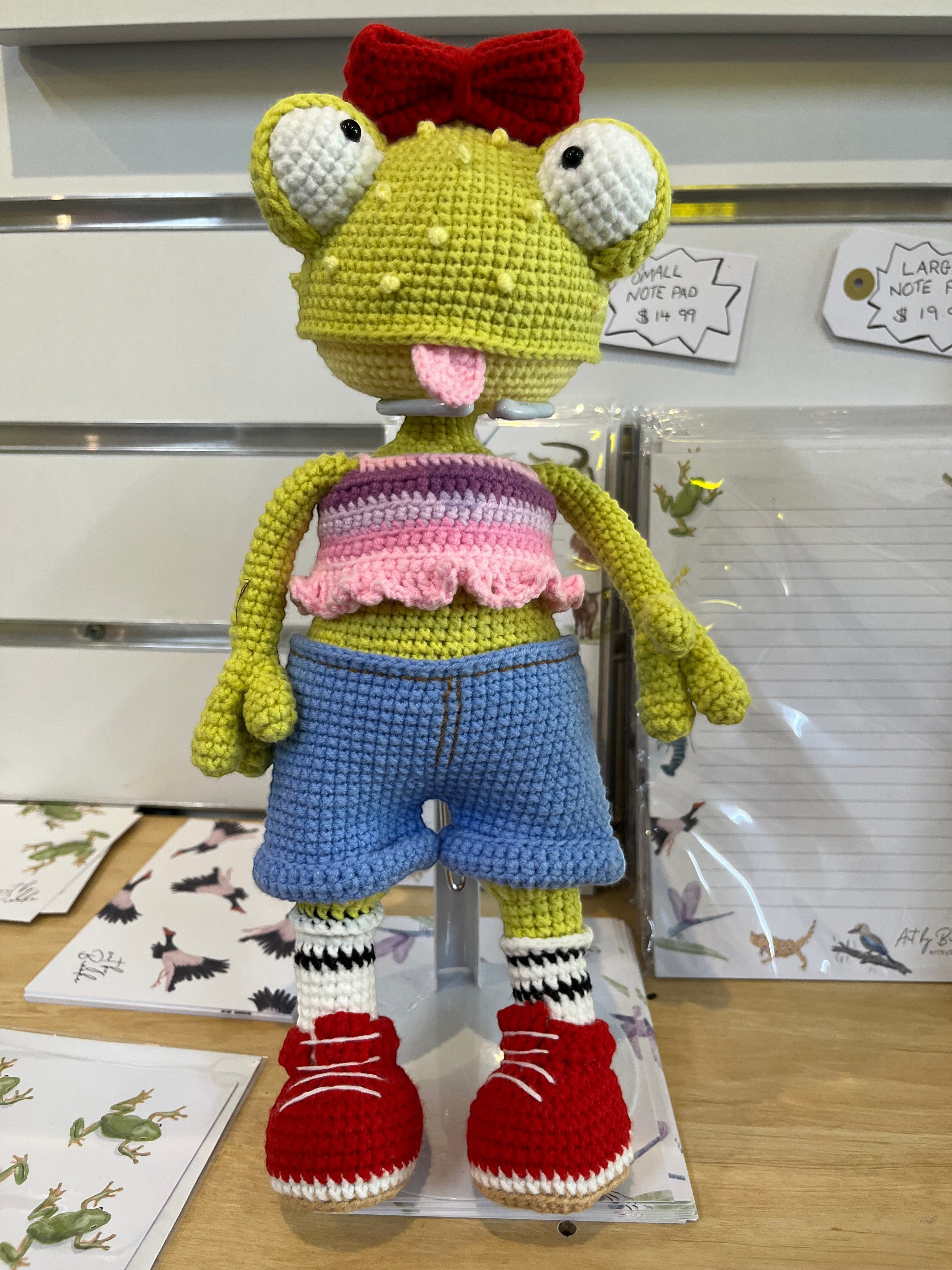 Suzie's Sunshine Dolls Frieda the Frog, a hand-crocheted doll with googly eyes, red bow, and tongue out. She wears a pastel crop top, denim shorts, striped socks, and red sneakers.