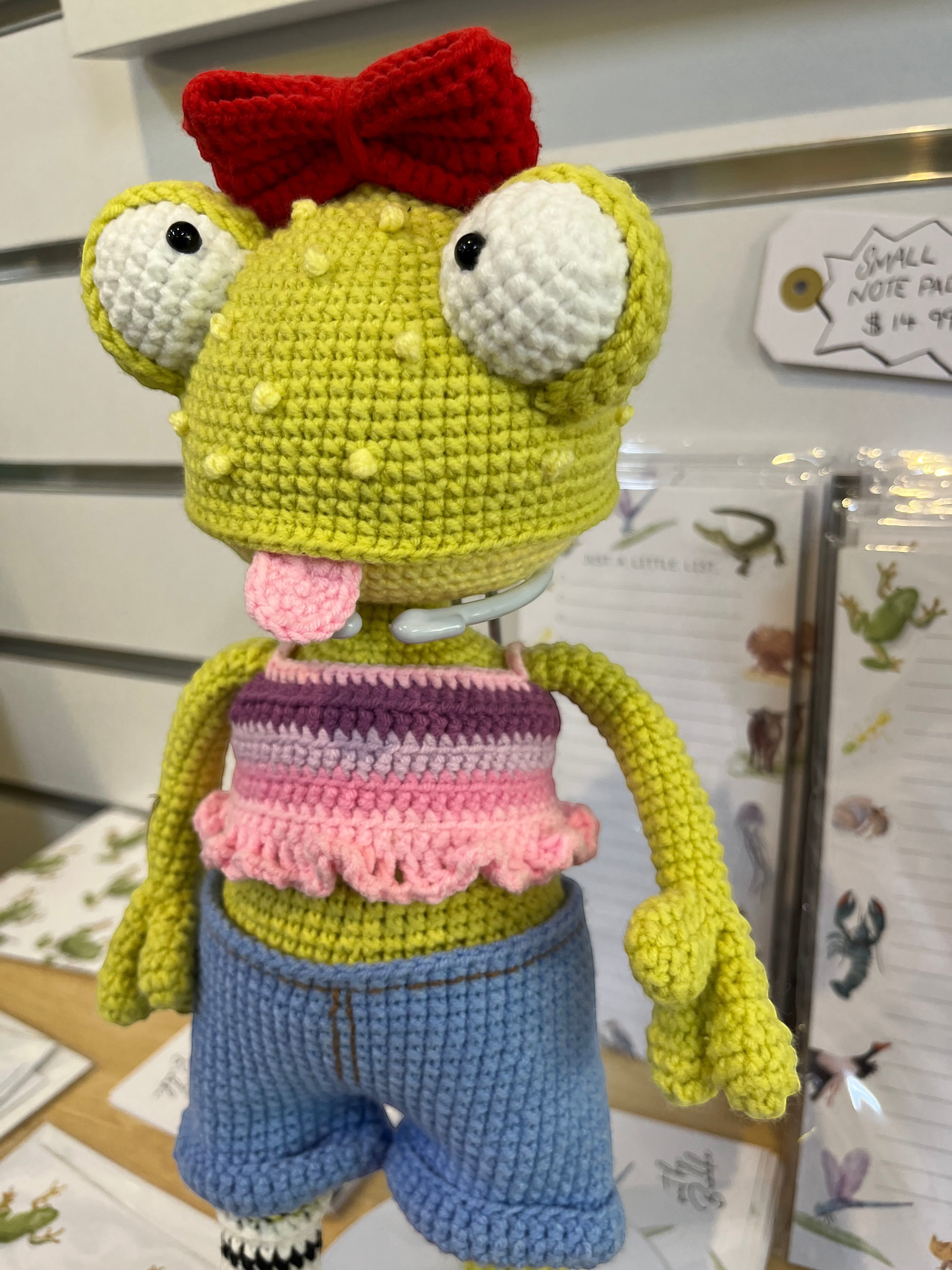 Suzie's Sunshine Dolls Frieda the Frog, a hand-crocheted doll with big googly eyes, red bow, and tongue out. She wears a pink and purple crop top and blue denim shorts.