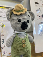 Suzie's Sunshine Dolls Kolby the Koala, a grey crocheted doll wearing green overalls and a straw hat.