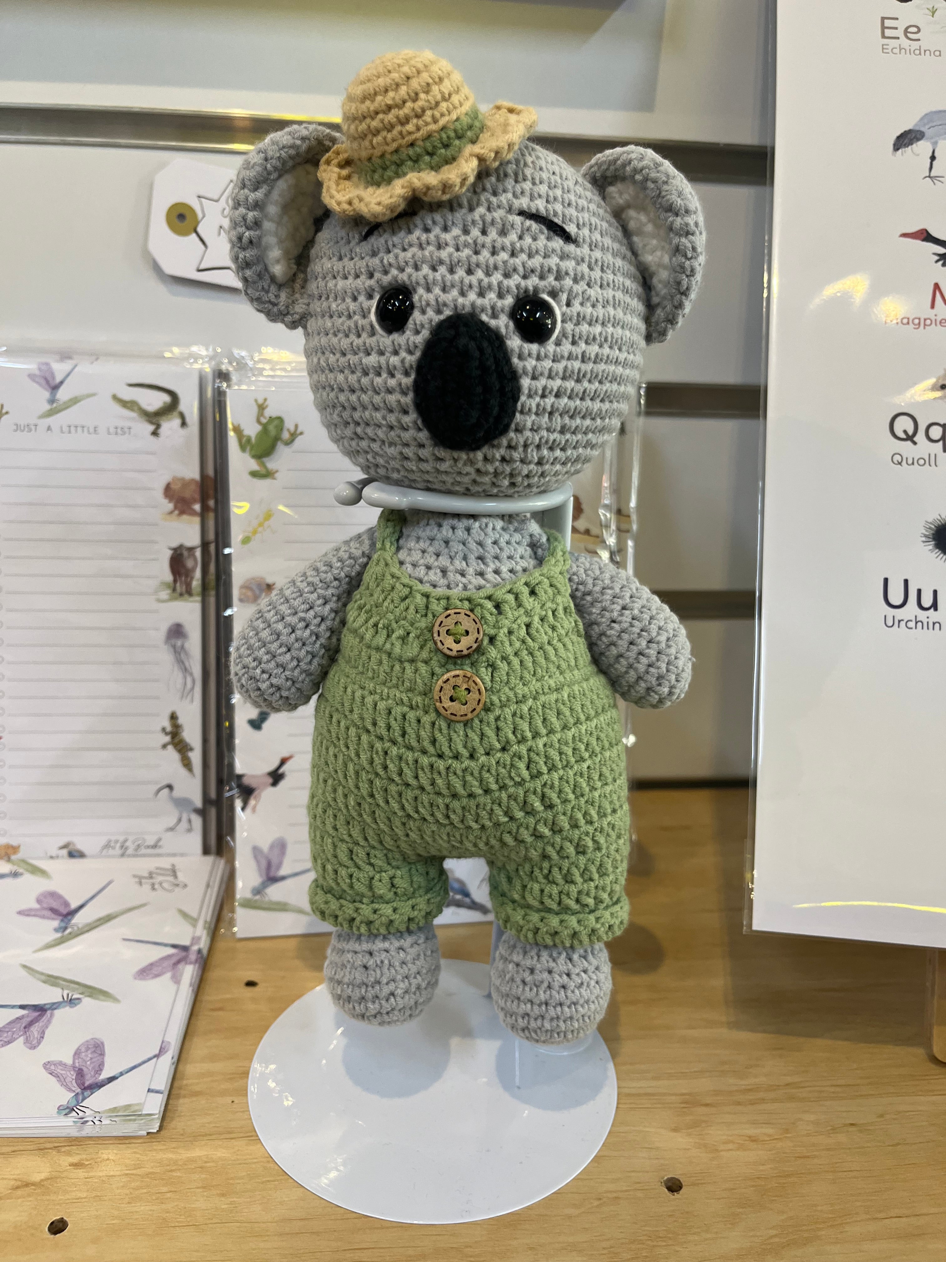Suzie's Sunshine Dolls Kolby the Koala, a grey crocheted stuffed animal wearing green overalls and a straw hat.
