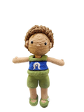 Suzie's Sunshine Dolls Malakai, a crocheted doll with short brown hair, wearing green shorts and a blue top with a white Dhari symbol.