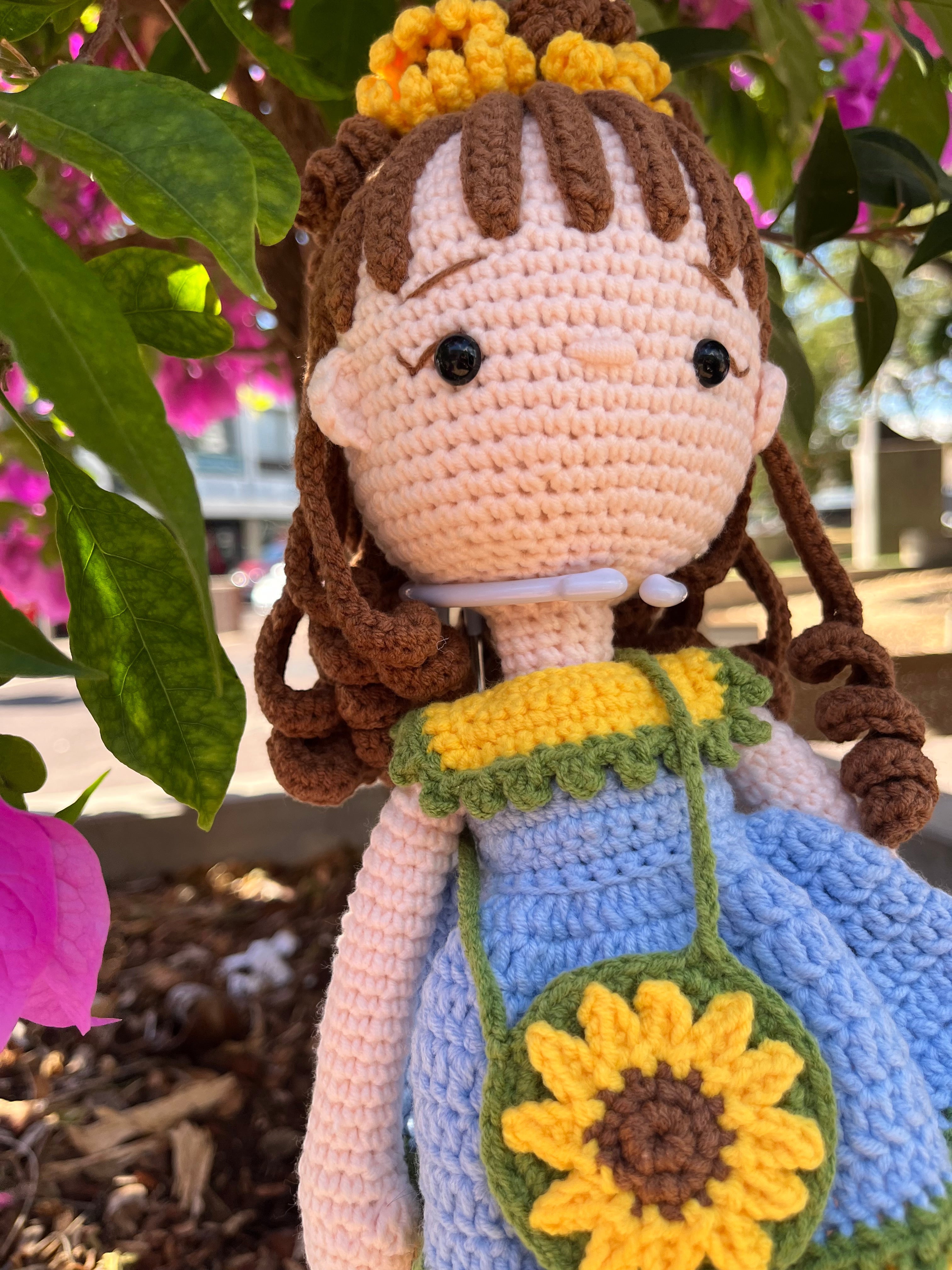 Suzie's Sunshine Dolls Sophie, a crocheted doll with brown yarn hair, wearing a blue dress with yellow trim and a sunflower purse. She has a yellow flower crown.