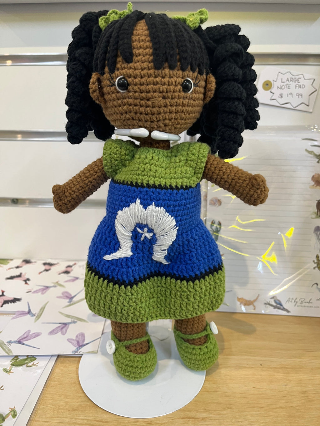 Suzie's Sunshine Dolls handmade crochet doll with long black curly hair, wearing a blue dress with green trim and a white Dhari symbol.